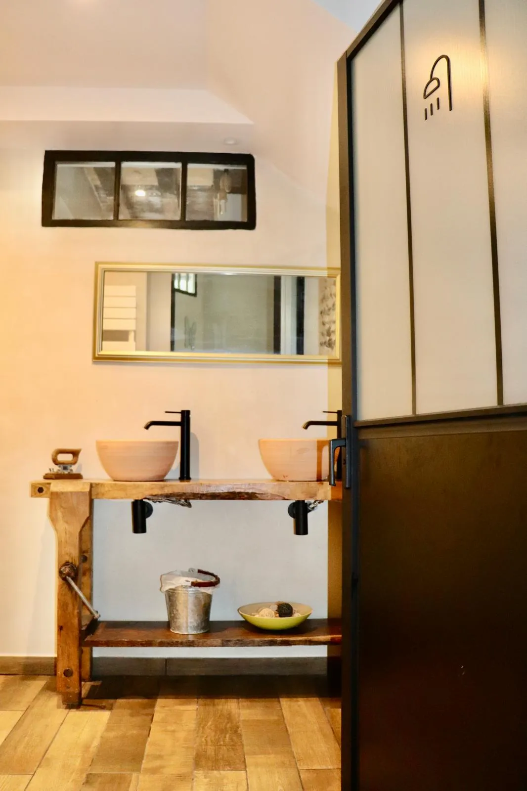 Salle de bain avec double vasque en céramique posée sur un établi ancien, à côté d’une verrière d’atelier