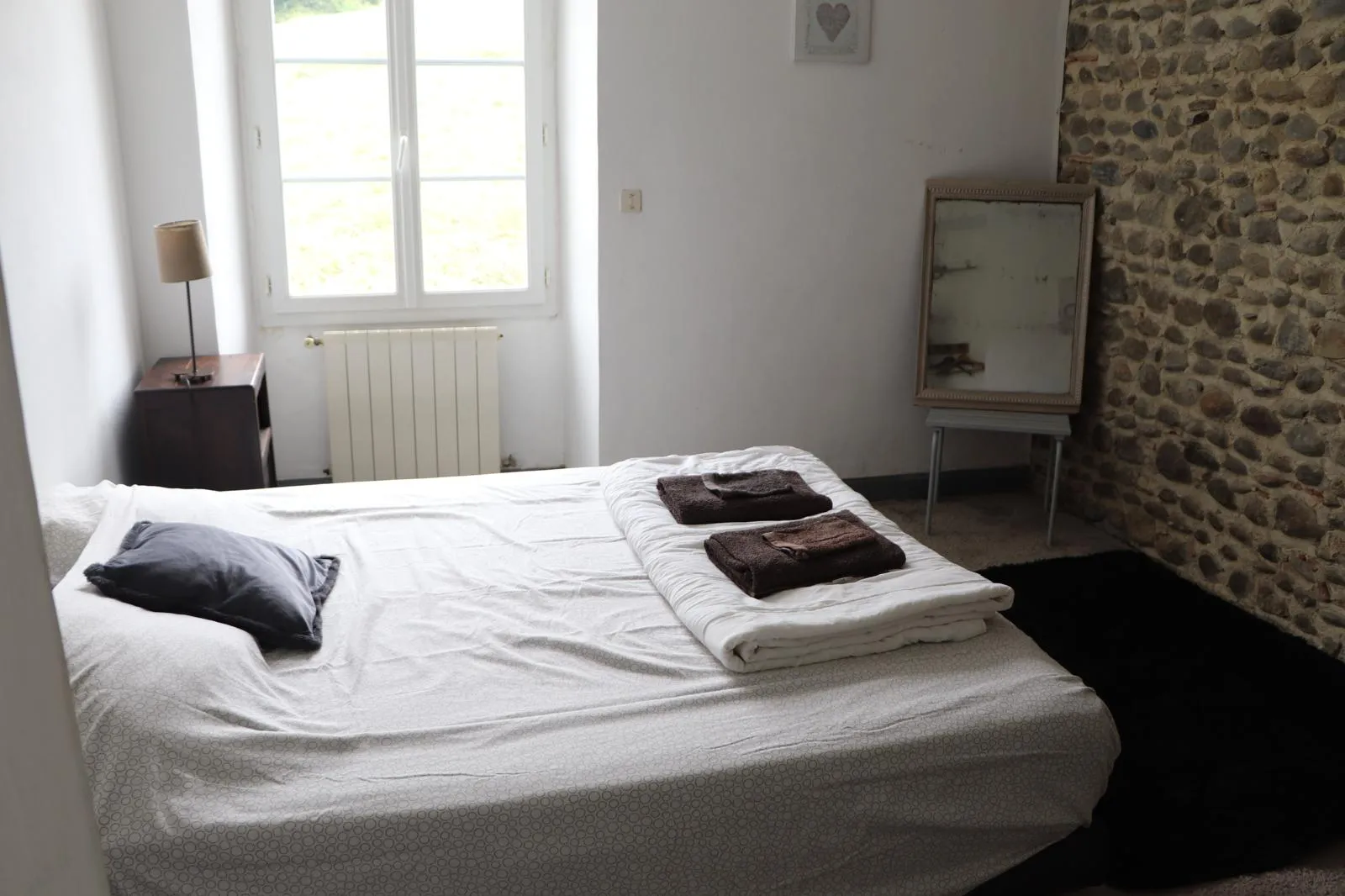 Chambre claire, lit double dressé, mur de galets et fenêtre à petits carreaux