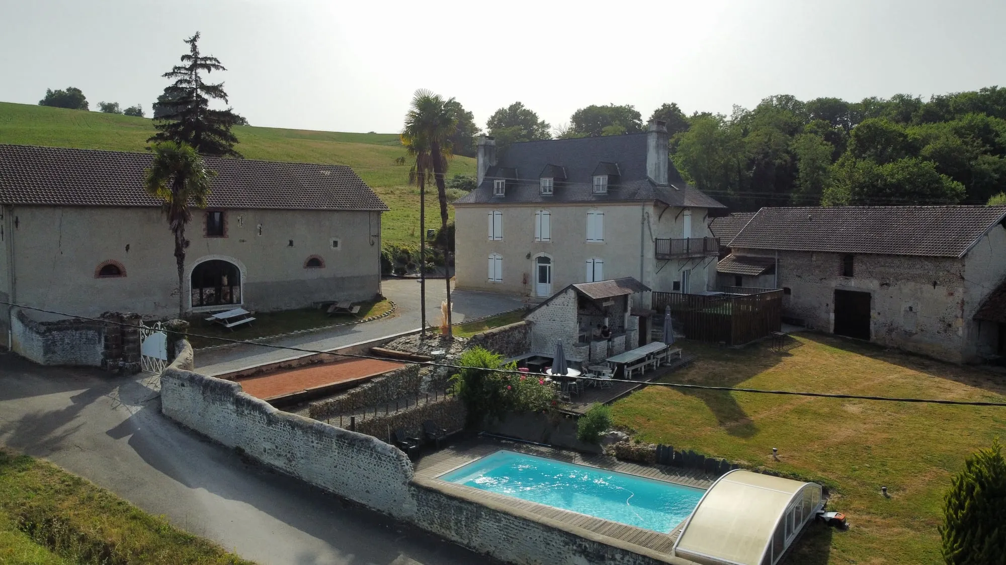 Vue aérienne des trois bâtiments alignés autour de la cour : la grange à arcade à gauche, la maison de maître à toit d’ardoise au centre, le long hangar de pierre à droite, la piscine en contrebas