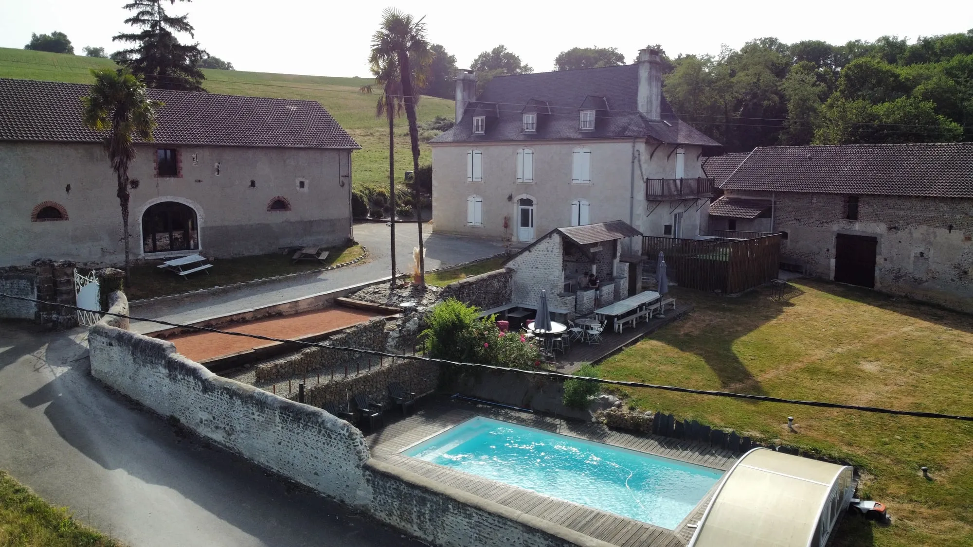 La piscine et son abri télescopique vus du ciel, la terrasse et ses parasols juste au-dessus, les bâtiments de la ferme à l’arrière-plan
