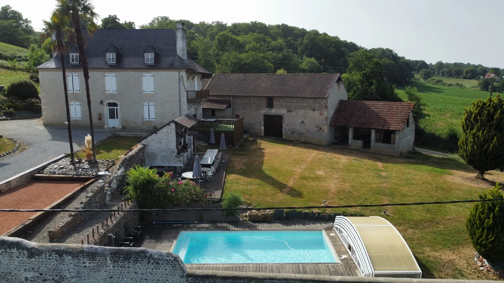 Vue aérienne depuis l’est : la maison de maître et ses palmiers à gauche, les granges en enfilade à droite, la grande pelouse au milieu et la piscine en bas