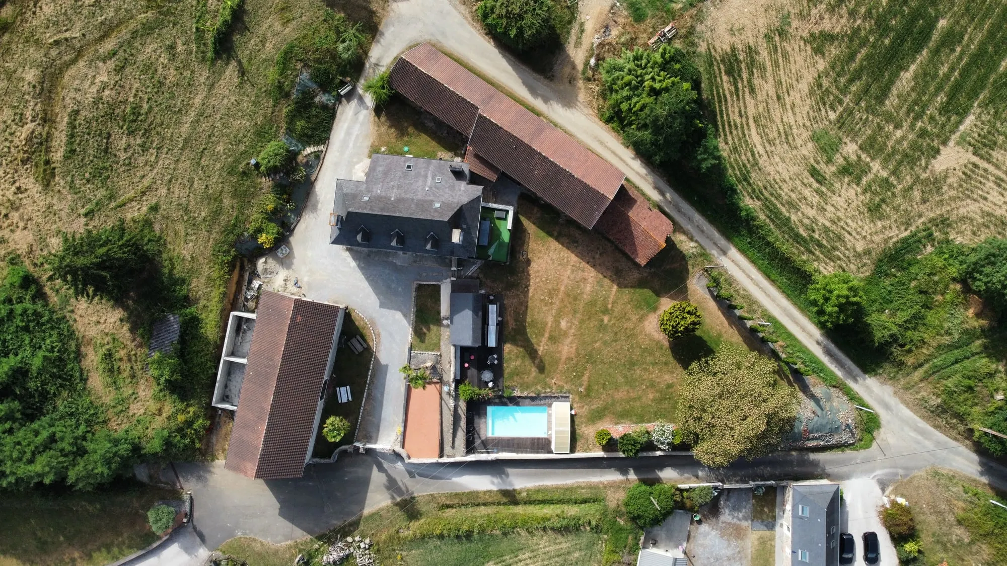 Vue à la verticale de la propriété : les toits de tuiles et d’ardoise, la cour gravillonnée, la piscine et la route qui longe les champs