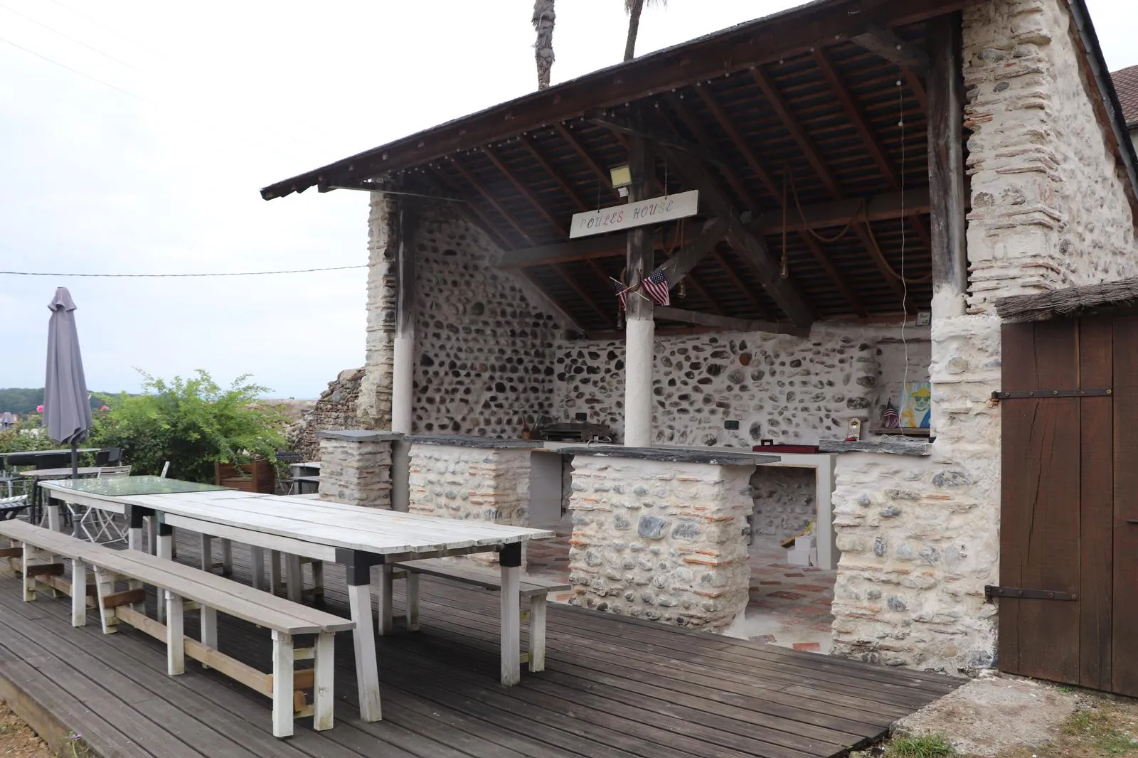 Terrasse couverte aux murs de galets, avec deux longues tables en bois et leurs bancs, ouverte sur la campagne