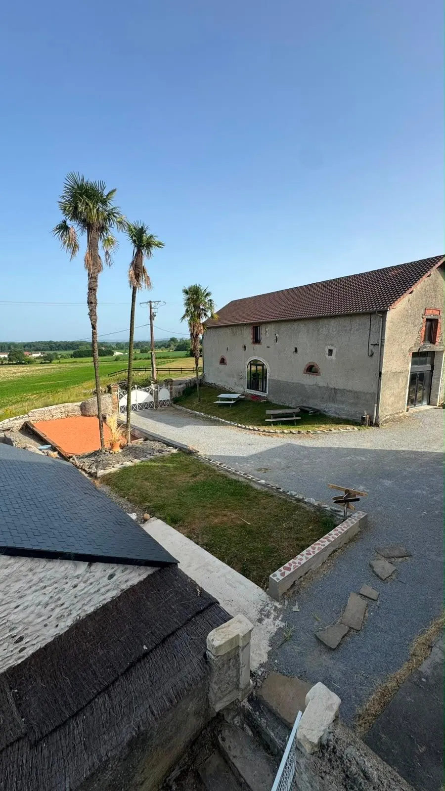 La cour gravillonnée et la grange en pierre sous un ciel bleu, palmiers au premier plan