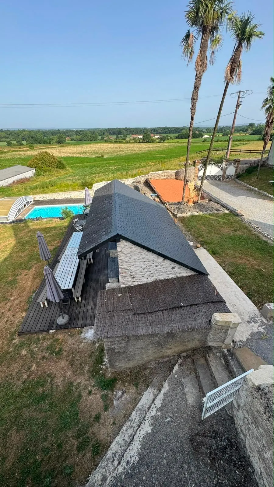 Vue plongeante sur le toit de la terrasse couverte, la piscine en contrebas et les champs jusqu’à l’horizon