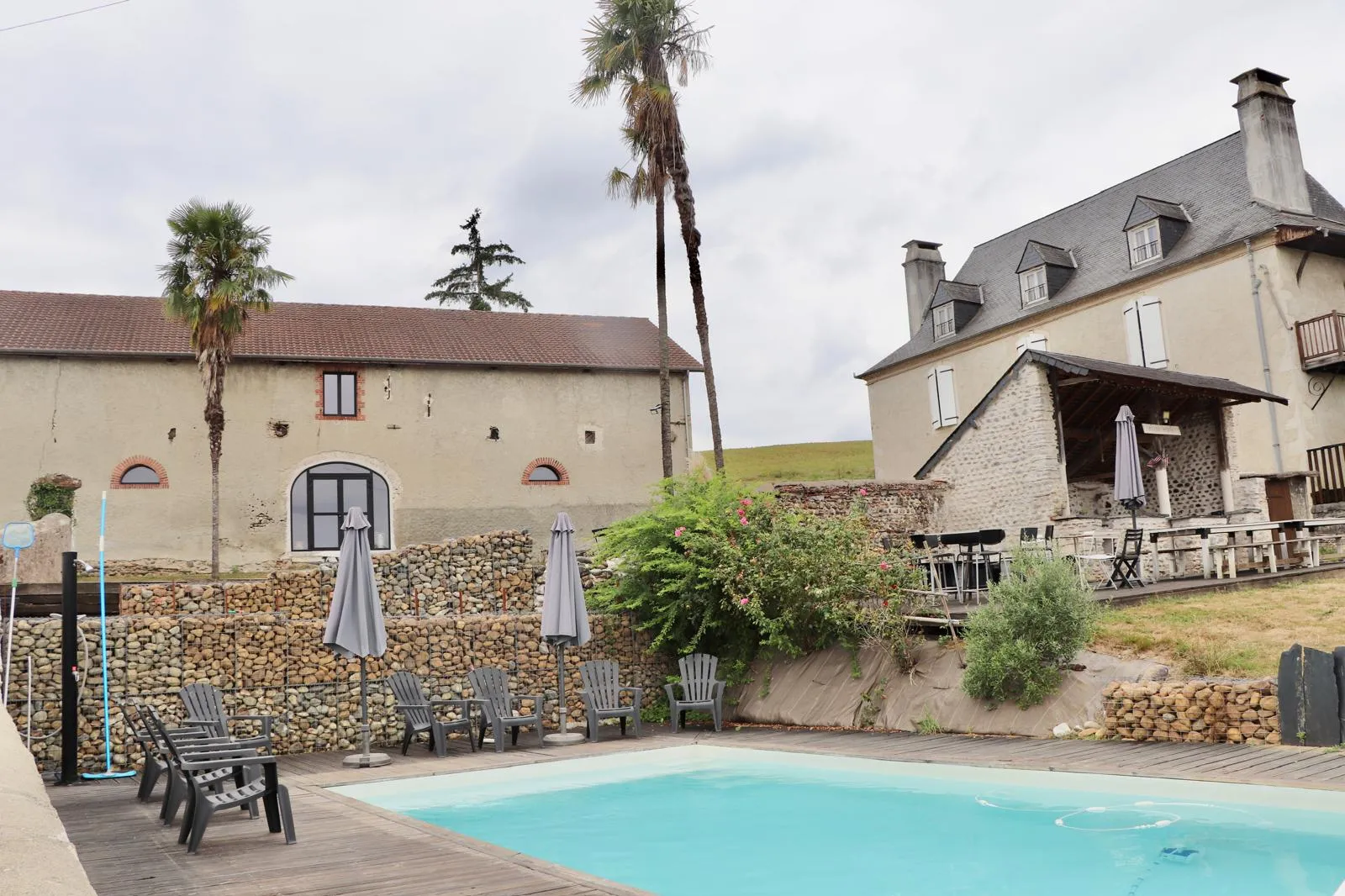La piscine et sa terrasse en bois, avec la maison béarnaise et la grange à l’arrière-plan