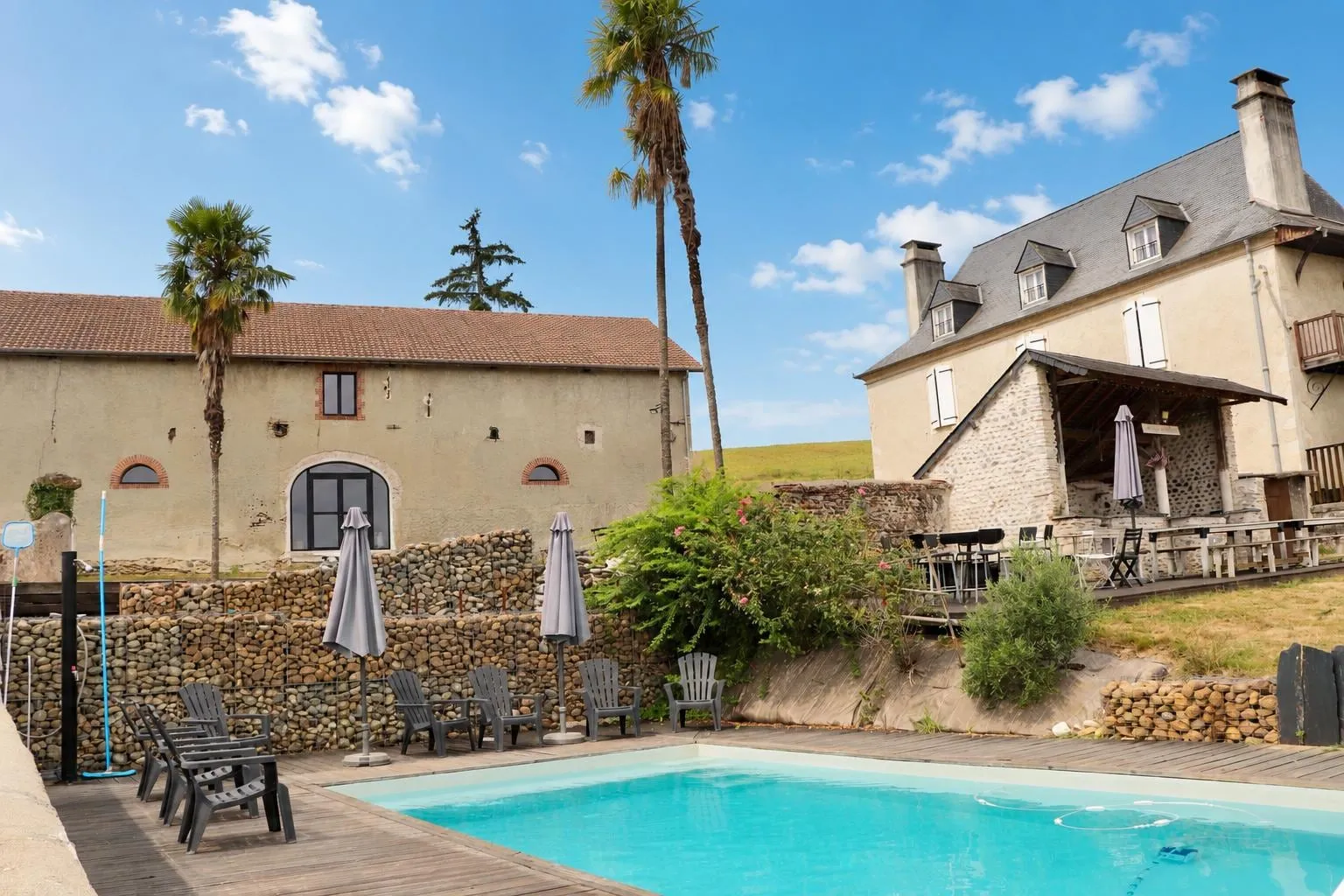 La piscine en plein soleil, transats et parasols repliés sur la terrasse en bois, la grange à gauche et la maison de maître à droite, palmiers se détachant sur un ciel bleu